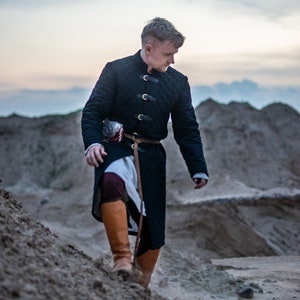 Peut inclure: Un homme portant une tunique matelass&eacute;e noire avec une ceinture et des bottes en cuir marron marche sur un chemin de sable.