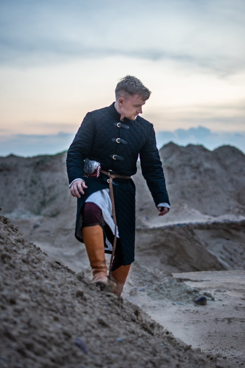Peut inclure: Un homme portant une tunique matelass&eacute;e noire avec une ceinture et des bottes en cuir marron marche sur un chemin de sable.
