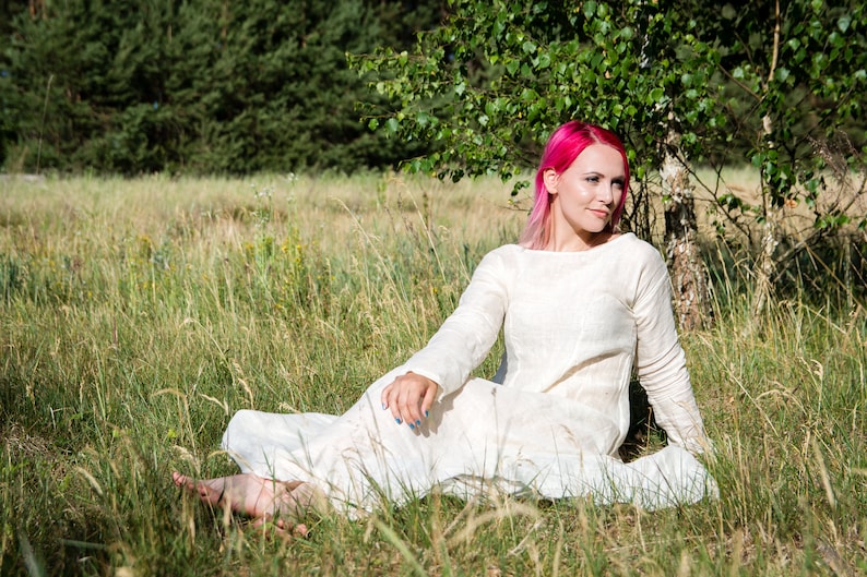 May include: A woman with pink hair sits in a field of tall grass wearing a white linen dress. She is barefoot and has her arms resting on her legs.
