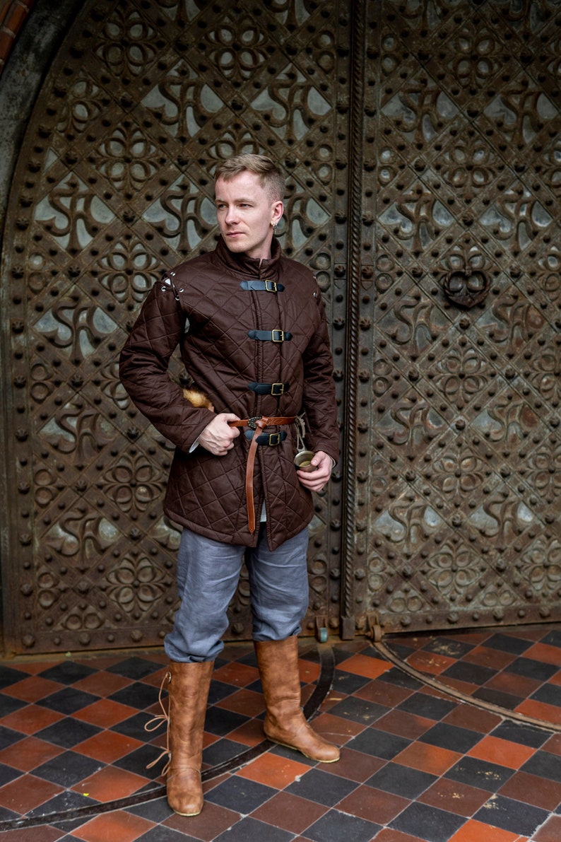 May include: A brown quilted jacket with black buckles and a brown leather belt. The jacket is paired with gray pants and brown leather boots. The background features a large, ornate metal door.
