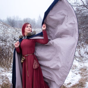 May include: A woman with red hair wearing a long, burgundy dress with a fur-trimmed, blue and gray hooded cloak. She is standing in a snowy field with trees in the background.