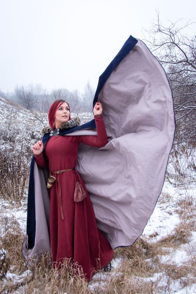 May include: A woman with red hair wearing a long, burgundy dress with a fur-trimmed, blue and gray hooded cloak. She is standing in a snowy field with trees in the background.