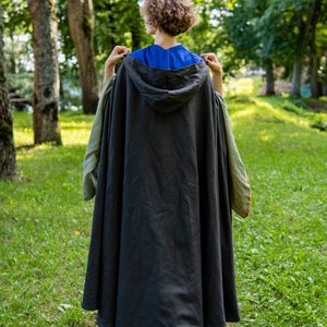 May include: A person wearing a dark gray hooded cloak with green and brown trim, standing in a grassy area with trees in the background.