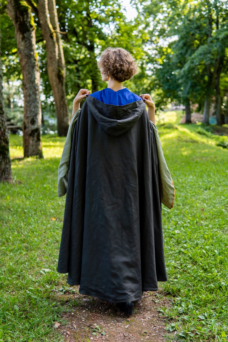 May include: A person wearing a dark gray hooded cloak with green and brown trim, standing in a grassy area with trees in the background.