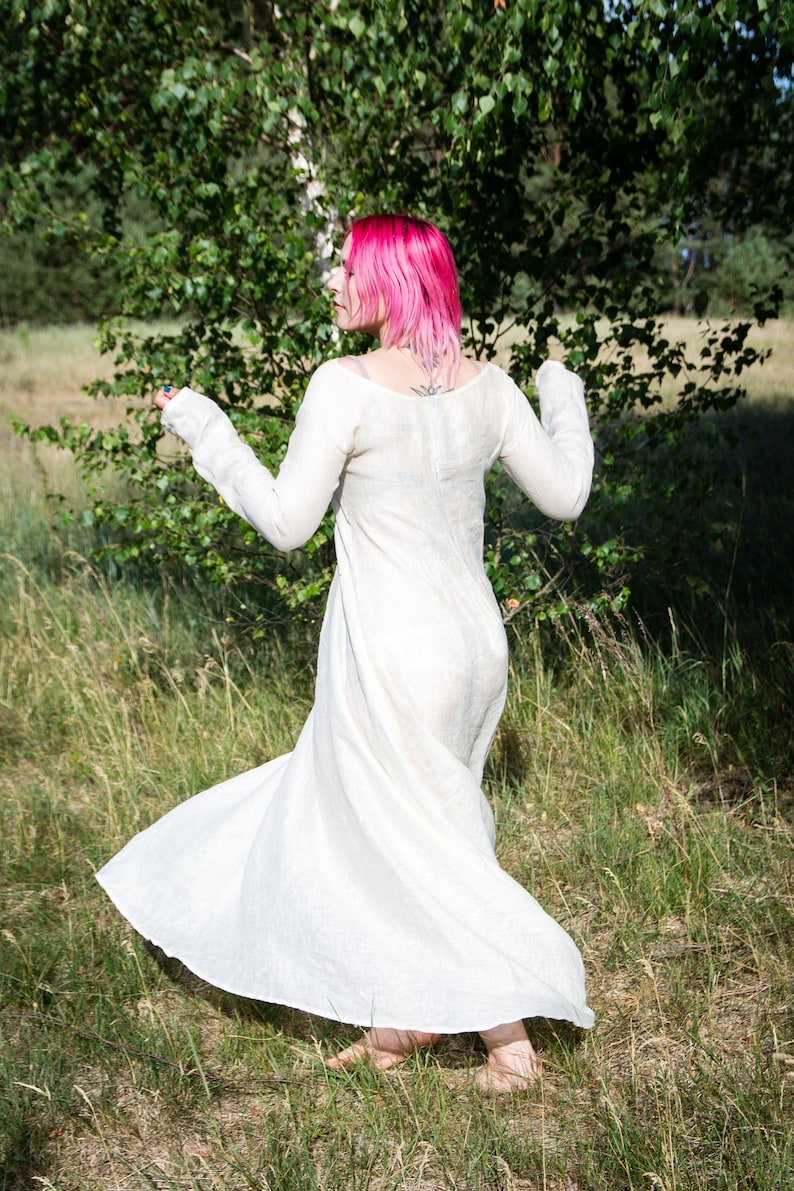 May include: A woman with pink hair wearing a white linen dress, standing in a field of tall grass.