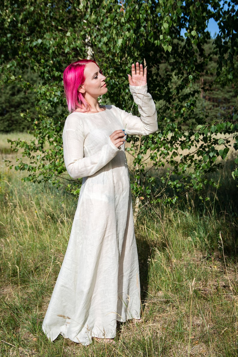 May include: A woman with pink hair wearing a long, white linen dress stands in a field of tall grass. She is holding a blade of grass in her hand and looking up at it.