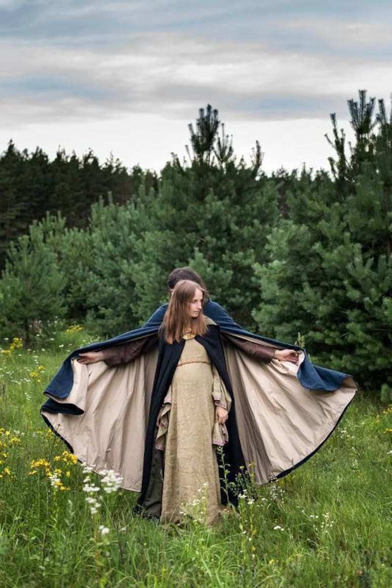 May include: A woman wearing a long, flowing, beige dress with a dark blue cape. The cape is lined with a lighter beige fabric and has a wide, flowing shape. The woman is standing in a field of green grass and wildflowers.