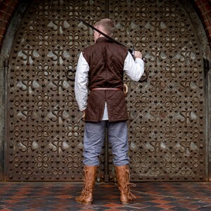 May include: A person wearing a brown quilted vest, white shirt, gray pants, and brown boots, holding a sword. The person is standing in front of a large, ornate, metal door. The background includes a brick archway.