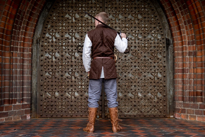 May include: A person wearing a brown quilted vest, white shirt, gray pants, and brown boots, holding a sword. The person is standing in front of a large, ornate, metal door. The background includes a brick archway.