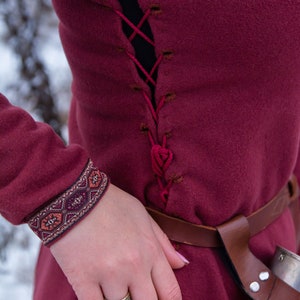 May include: A burgundy tunic with a laced-up back, a brown leather belt, and a decorative cuff. The tunic is worn with a ring on the index finger.