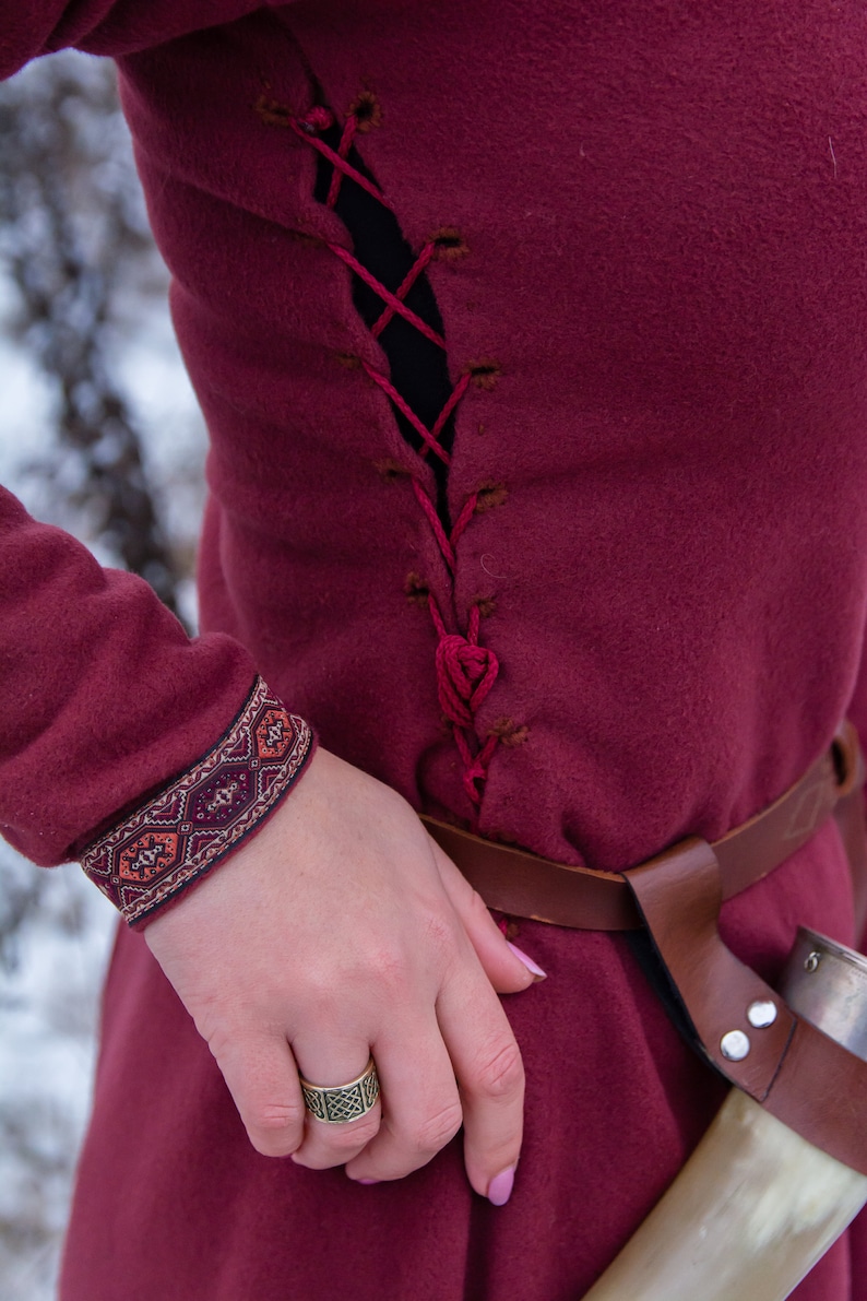 May include: A burgundy tunic with a laced-up back, a brown leather belt, and a decorative cuff. The tunic is worn with a ring on the index finger.