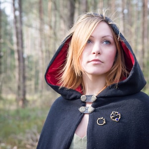 May include: A woman wearing a black hooded cloak with a red lining. The cloak has decorative metal buttons and a brooch with blue gemstones. She is standing in a forest.