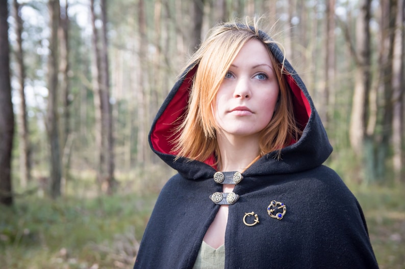 May include: A woman wearing a black hooded cloak with a red lining. The cloak has decorative metal buttons and a brooch with blue gemstones. She is standing in a forest.