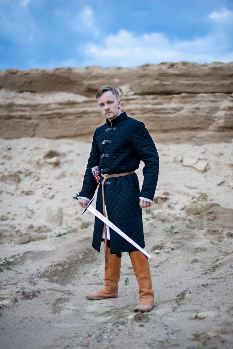 Peut inclure: Un homme v&ecirc;tu d'un manteau matelass&eacute; noir avec une ceinture marron et des bottes marron tient une &eacute;p&eacute;e argent&eacute;e devant lui. Il se tient dans une zone sablonneuse avec une falaise rocheuse en arri&egrave;re-plan.