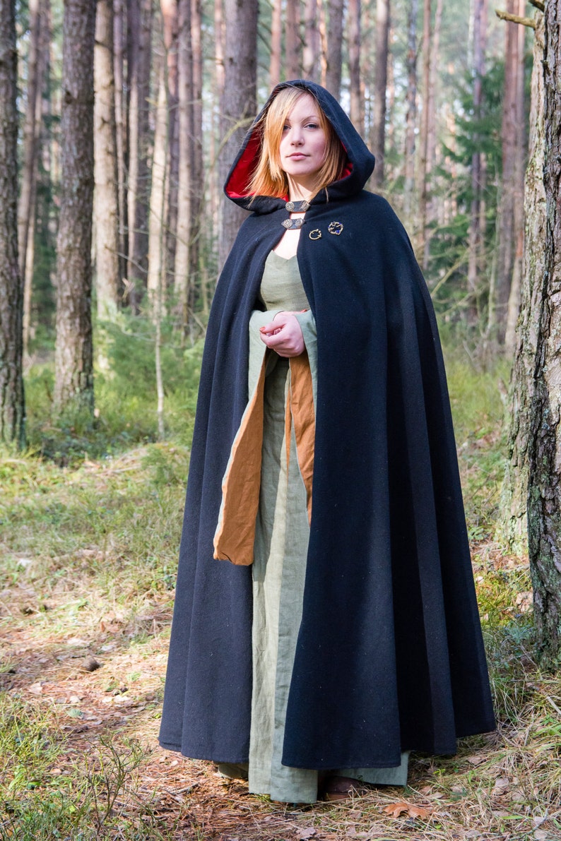May include: A woman wearing a black hooded cloak with a red lining, standing in a forest. The cloak has a decorative buckle and is worn over a green dress.