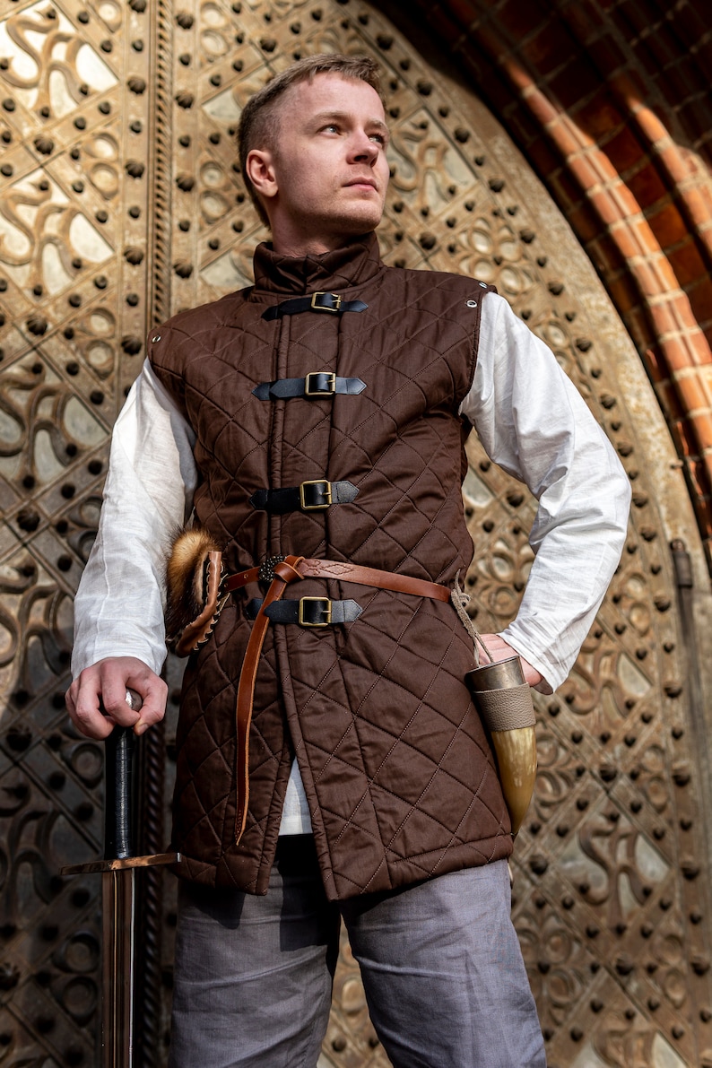 May include: A brown quilted vest with black buckles and a leather belt. The vest is worn over a white shirt and paired with gray pants. A sword and a horn are also visible.
