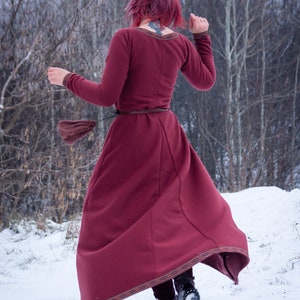 May include: A woman in a long, burgundy dress with a brown belt walks through a snowy forest. The dress has a decorative trim around the hem.