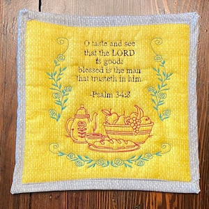 May include: A yellow fabric kitchen pot holder with a gray border. It features an embroidered design of a teapot, a bowl of fruit, and bread. The text reads "O taste and see that the LORD is good: blessed is the man that trusteth in him. -Psalm 34:8"