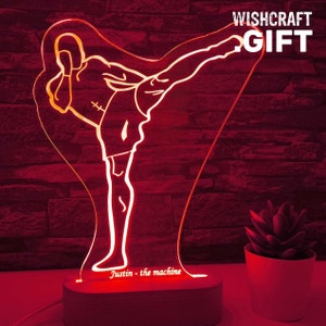 Peut inclure: Une veilleuse LED rouge avec une silhouette d'une personne donnant un coup de pied en position de combat d'arts martiaux. La lumière a une base en bois et le texte "Justin - the machine" est imprimé sur la base.