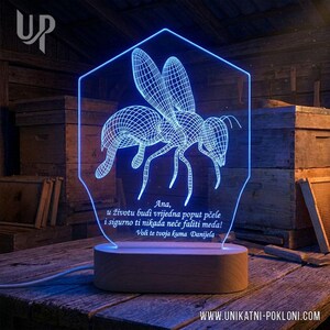 Pode incluir: Lâmpada LED azul com um design de abelha em arame dentro de uma moldura hexagonal. A lâmpada está sobre uma base de madeira e contém texto em língua estrangeira. O endereço do site é visível na parte inferior.