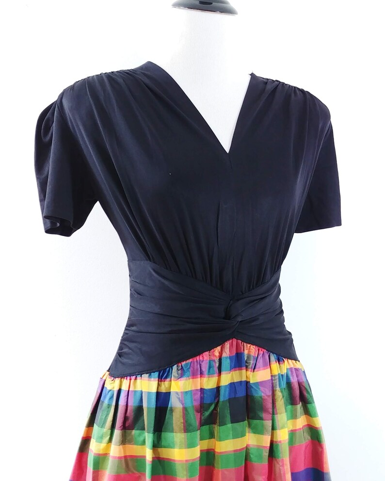 May include: A black short-sleeved dress with a V-neckline and a fitted bodice. The skirt is a colorful plaid pattern with red, yellow, green, blue, and pink.