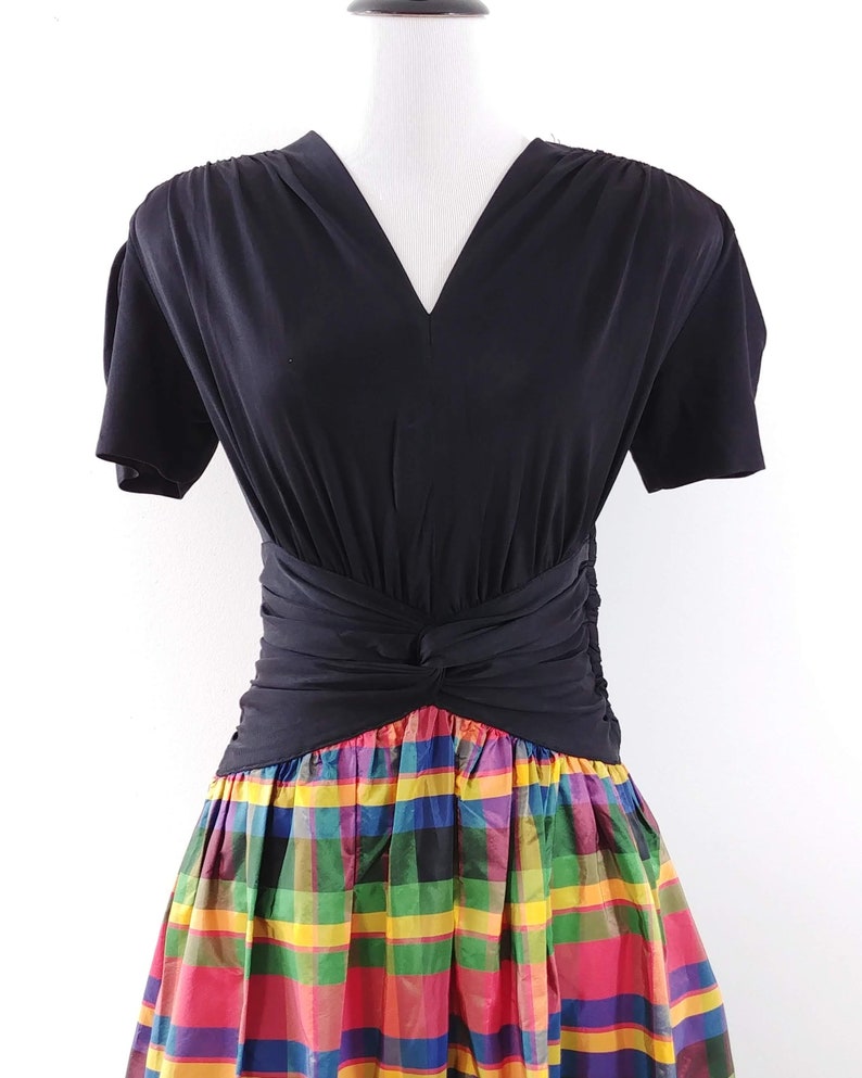 May include: A black short-sleeved dress with a V-neckline and a knotted waistband. The skirt is a colorful plaid pattern with red, yellow, green, blue, and purple.