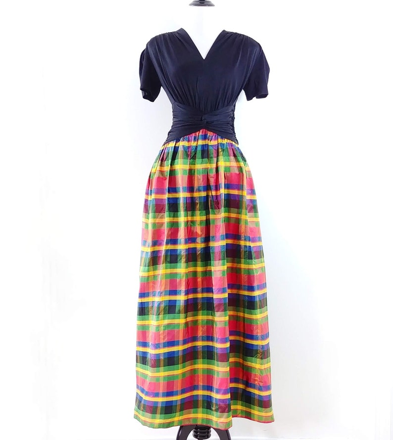 May include: A black short-sleeved top with a V-neckline and a knot at the waist, paired with a long, colorful plaid skirt. The skirt features a red, yellow, green, blue, and black plaid pattern.