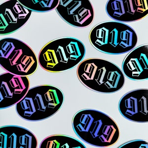 Op de afbeelding: Een verzameling van 15 ovale stickers met het nummer 919 gedrukt in een gotisch lettertype. De stickers zijn iriserend en hebben een regenboogachtige glans.
