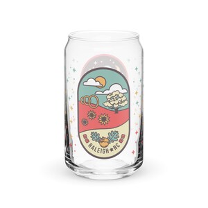 Può includere: Una lattina di vetro trasparente con un design colorato che presenta un albero, il sole, nuvole e girasoli. Il design include anche il testo "RALEIGH * NC" e una piccola ghianda.