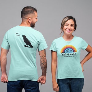 May include: Two light blue t-shirts with different designs. The first shirt has a black crow with the text "Caw Caw Caw" above it. The second shirt has a rainbow and mountains with the text "The aisle of shame is camping and I went too".