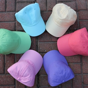 Puede incluir: Seis gorras de béisbol lisas en diferentes colores pastel: azul claro, beige, rosa, verde, morado y coral. Las gorras están dispuestas en círculo sobre una superficie de ladrillo.