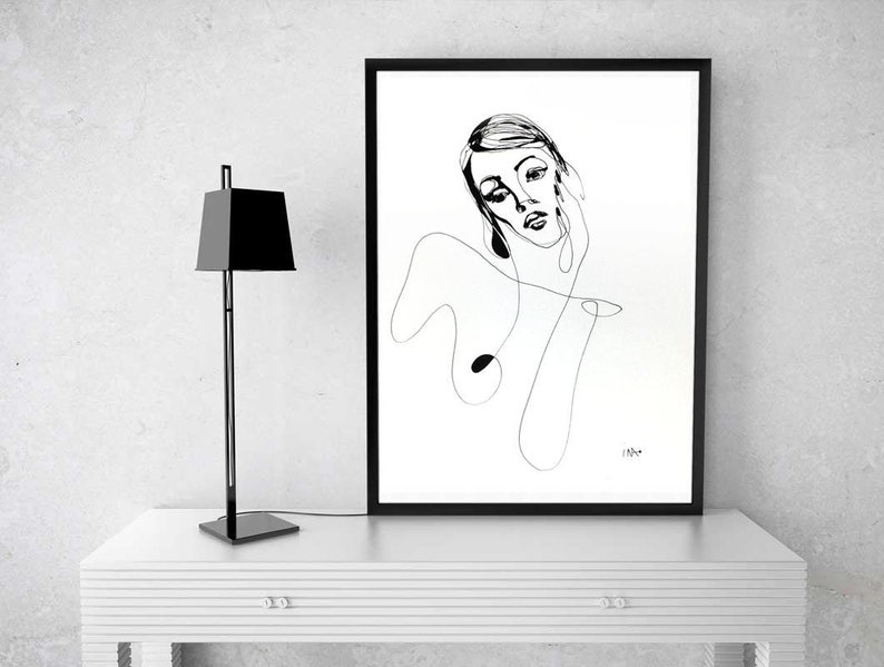 Może przedstawiać: Czarny obraz w ramie z minimalistycznym rysunkiem linii twarzy i g&oacute;rnej części ciała kobiety. Czarna, nowoczesna lampa stoi po lewej stronie grafiki na białym stole. Grafika to prosty, elegancki design.