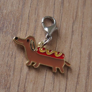 Puede incluir: Un charm de dachshund dorado con un perrito caliente rojo y amarillo en la espalda. El charm tiene un lazo de metal plateado para sujetarlo a una bolsa o llavero.
