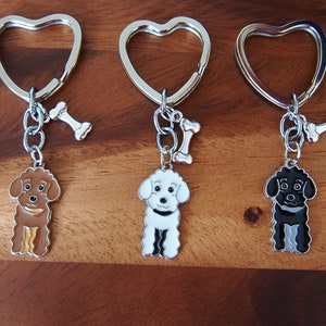 May include: Three heart-shaped keyrings with silver-tone metal rings and charms. Each charm features a cartoon poodle in brown, white, and black, with a bone charm attached.