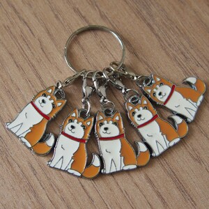 May include: A keychain with six metal charms featuring cartoon Shiba Inu dogs. The dogs are orange and white with red collars. The charms are attached to a silver key ring.