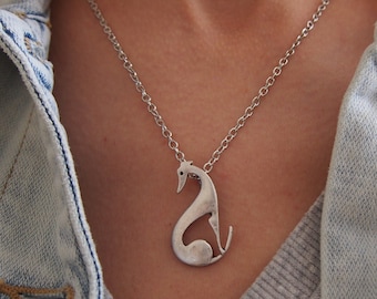 Puede incluir: Un collar con colgante plateado con un diseño estilizado de galgo. El colgante de perro cuelga de una delicada cadena plateada. El collar se lleva sobre un suéter gris y una chaqueta vaquera azul claro.