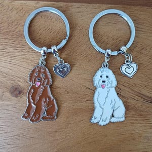 May include: Two dog keyrings. One has a brown dog charm, the other a white dog charm. Each charm is attached to a silver keyring and a heart-shaped charm. The keyrings are displayed on a wooden surface.