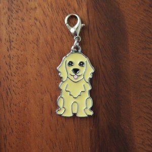 Könnte beinhalten: Ein goldfarbener Metallanhänger mit einem sitzenden Golden Retriever mit herausgestreckter rosa Zunge. Der Anhänger hat eine silberne Metallschaufel zum Anbringen an einem Schlüsselbund oder einer Tasche.