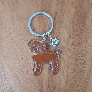 May include: A brown poodle keychain with a silver ring and a silver heart charm. The poodle is standing on all four legs with its tongue sticking out.
