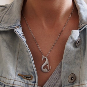 May include: A silver-colored pendant necklace featuring a stylized greyhound dog design. The dog is depicted in a seated position with its head and tail raised. The necklace is worn over a light blue denim jacket.