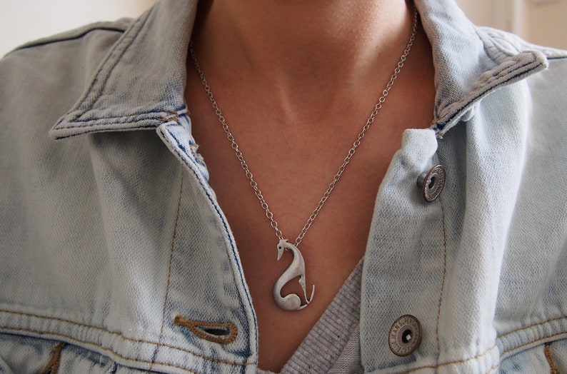May include: A silver-colored pendant necklace featuring a stylized greyhound dog design. The dog is depicted in a seated position with its head and tail raised. The necklace is worn over a light blue denim jacket.