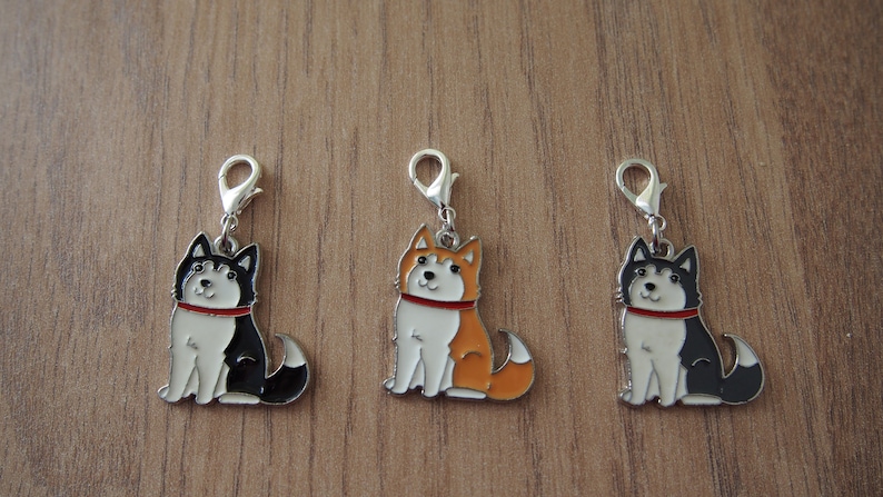 May include: Three metal charms featuring cartoon husky dogs. The dogs are sitting and have red collars. The charms are silver and have a clip on the top. The dogs are black and white, orange and white, and black and white.