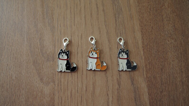 May include: Three metal charms featuring cartoon husky dogs in black, white, and orange. Each charm has a silver loop for attaching to a keychain or bag.