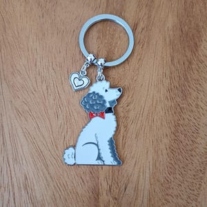 May include: A silver key chain with a white and gray poodle charm and a silver heart charm. The poodle is wearing a red bow tie.