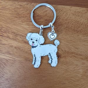 May include: A silver keychain featuring a white poodle charm and a silver heart charm. The poodle charm has black eyes and a pink nose.