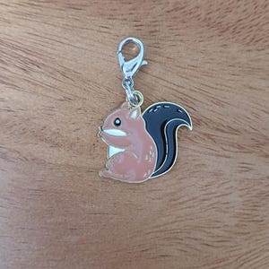 May include: A silver charm with a brown squirrel with a black tail. The squirrel is holding a nut in its paws.