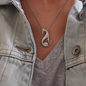 May include: A silver-toned pendant necklace featuring a stylized greyhound dog design. The pendant hangs from a delicate chain and is worn over a grey shirt and a light blue denim jacket.