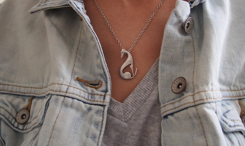 May include: A silver-toned pendant necklace featuring a stylized greyhound dog design. The pendant hangs from a delicate chain and is worn over a grey shirt and a light blue denim jacket.