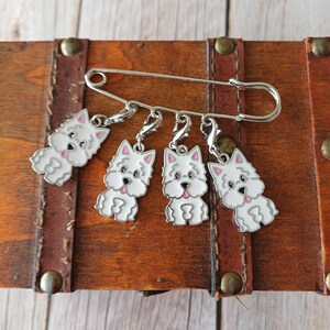 May include: A silver-toned metal safety pin featuring four dangling charms, each shaped like a white West Highland White Terrier dog with pink-accented ears and tongue, attached by small lobster claw clasps to the main pin structure.