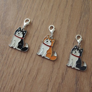 May include: Three metal charms featuring cartoon husky dogs in different colors: black and white, orange and white, and black and white. The charms have a silver finish and a loop for attaching to a keychain or bag.